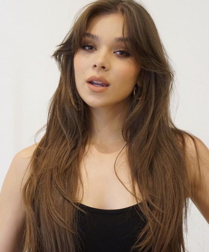 a woman with long brown hair wearing a black top and posing for the camera in front of a white wall