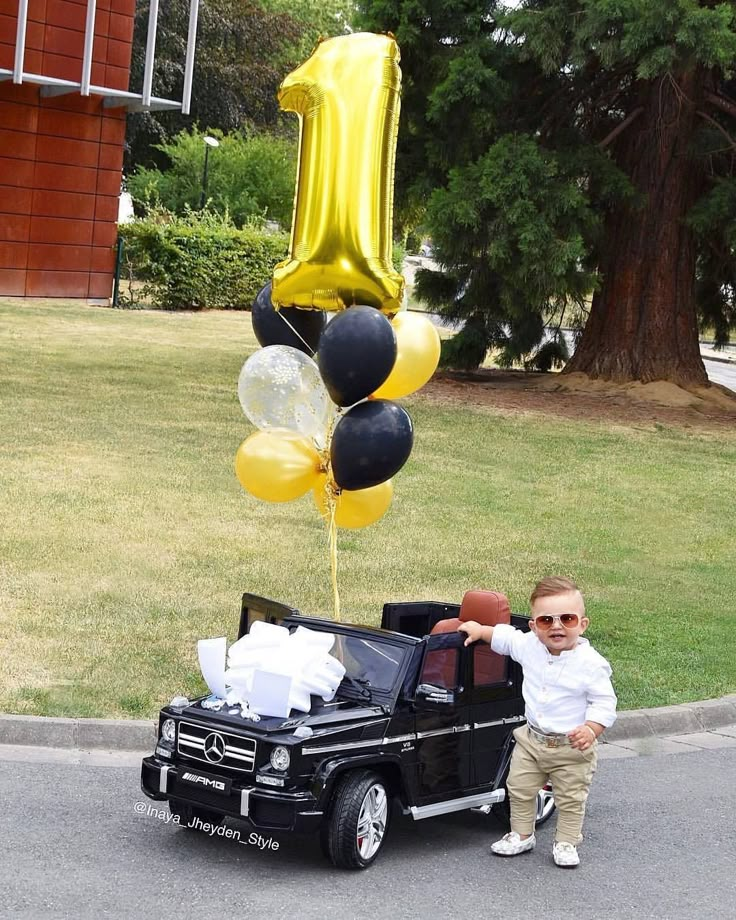 a little boy standing next to a black car with balloons in the shape of number one