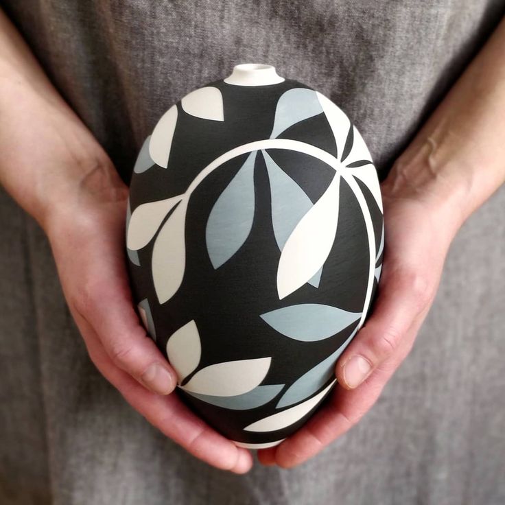 a person holding a black and white vase in their hands with leaves painted on it