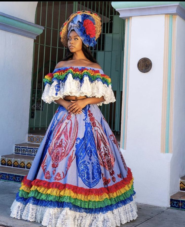 a woman in a colorful dress and hat standing on the side walk with her hands on her hips