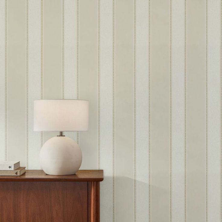 a white lamp sitting on top of a wooden dresser next to a wallpapered wall