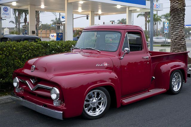 an old red pickup truck parked in front of a gas station with palm trees behind it