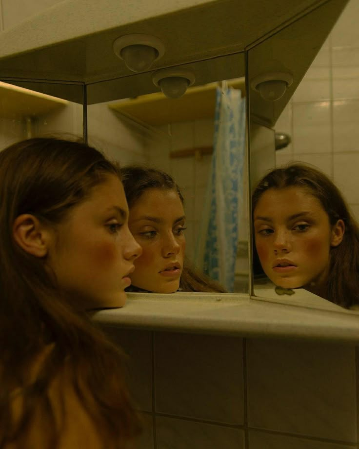 three girls are looking in the mirror and one girl is staring at her own reflection