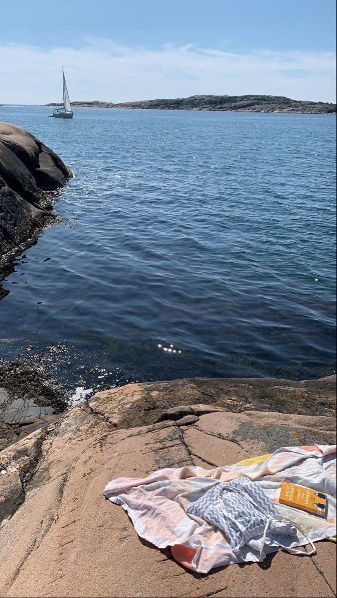 there is a blanket on the rocks by the water with a sailboat in the distance