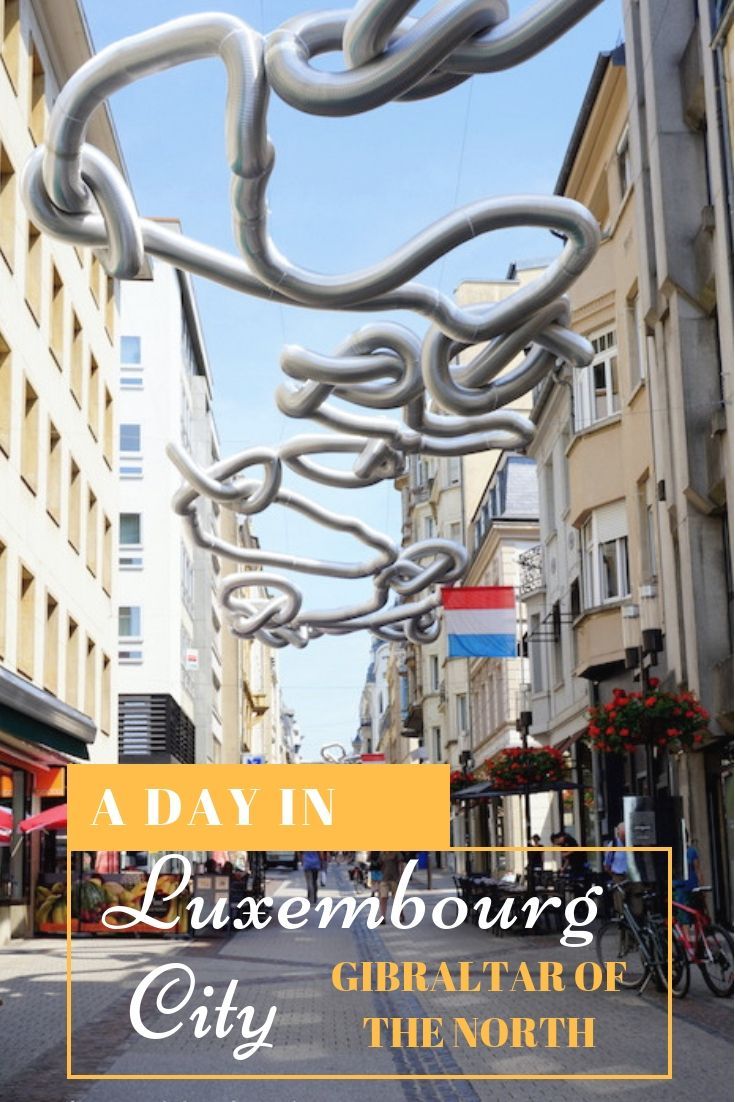 a city street with buildings and people walking on the side walk that has a large metal sculpture hanging from it's sides