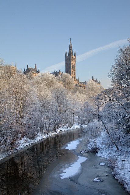 a river running through a snow covered forest next to a tall building with a clock tower in the background