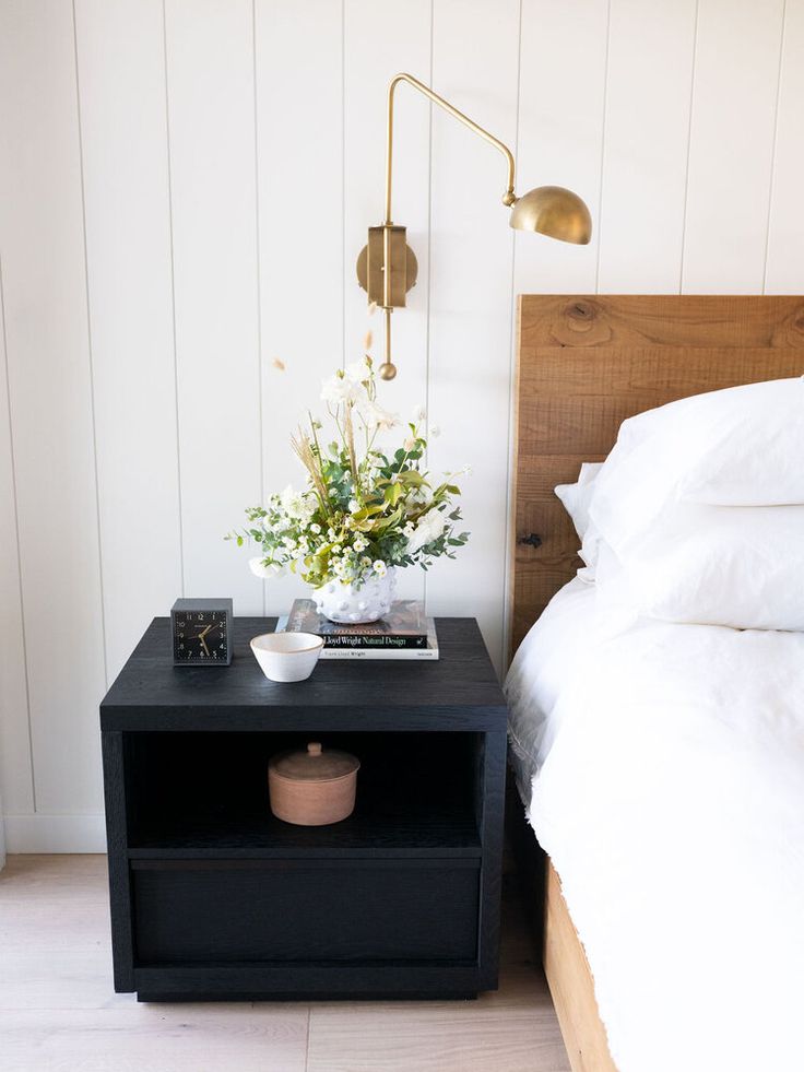 a nightstand with flowers on it next to a bed in a white walled room and wood paneled walls