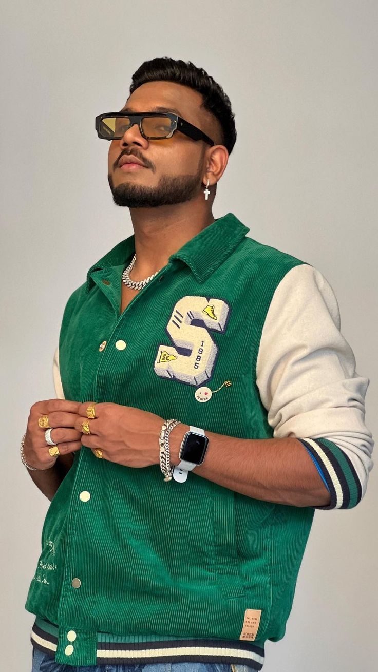 a man wearing a green vest and gold ring with his hand on his hip while standing in front of a gray background