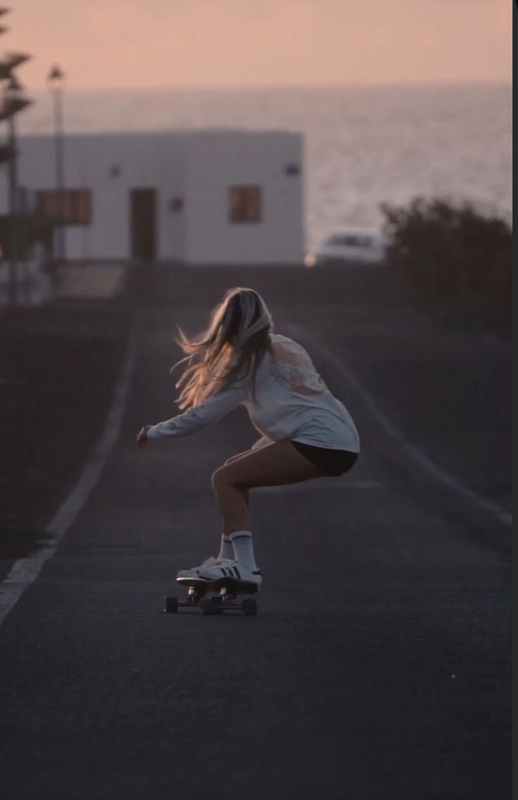 a woman riding a skateboard down a street next to the ocean at sunset or dawn