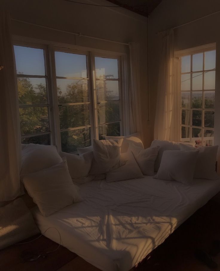 a bed sitting in front of two windows with white pillows on top of it and the sun shining through them