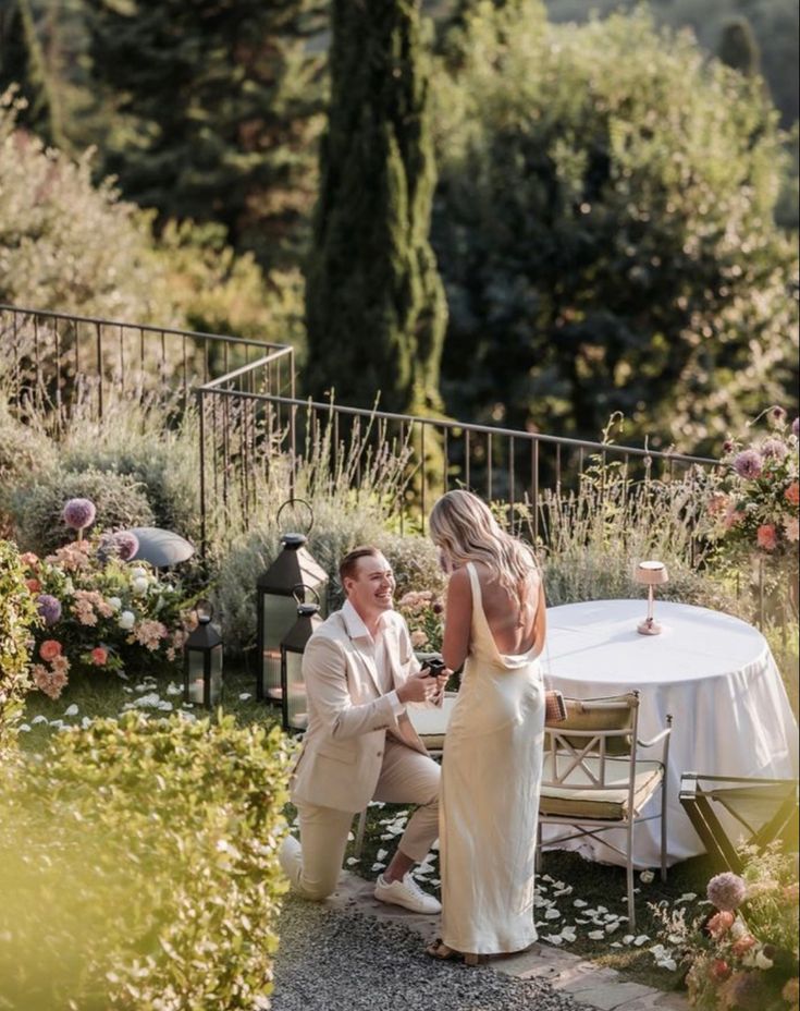 a man and woman sitting at a table in the middle of a garden with flowers