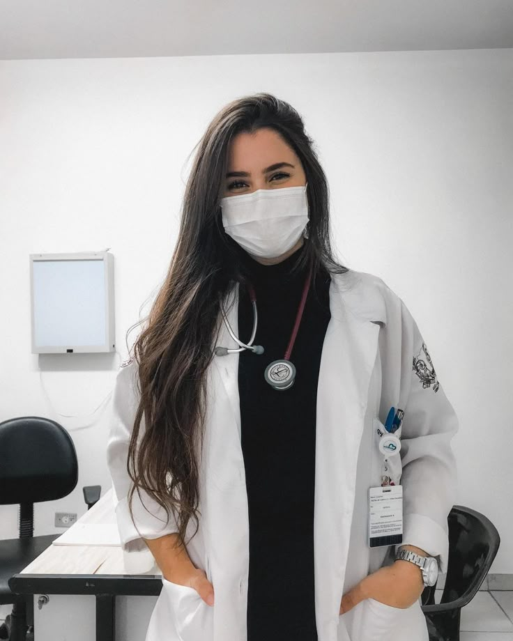 a woman wearing a white lab coat and medical mask standing in front of a desk