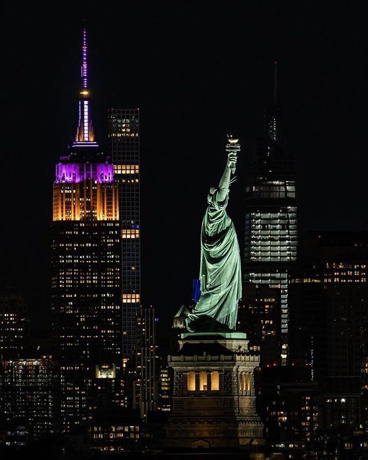 the statue of liberty lit up at night in new york city, with skyscrapers behind it