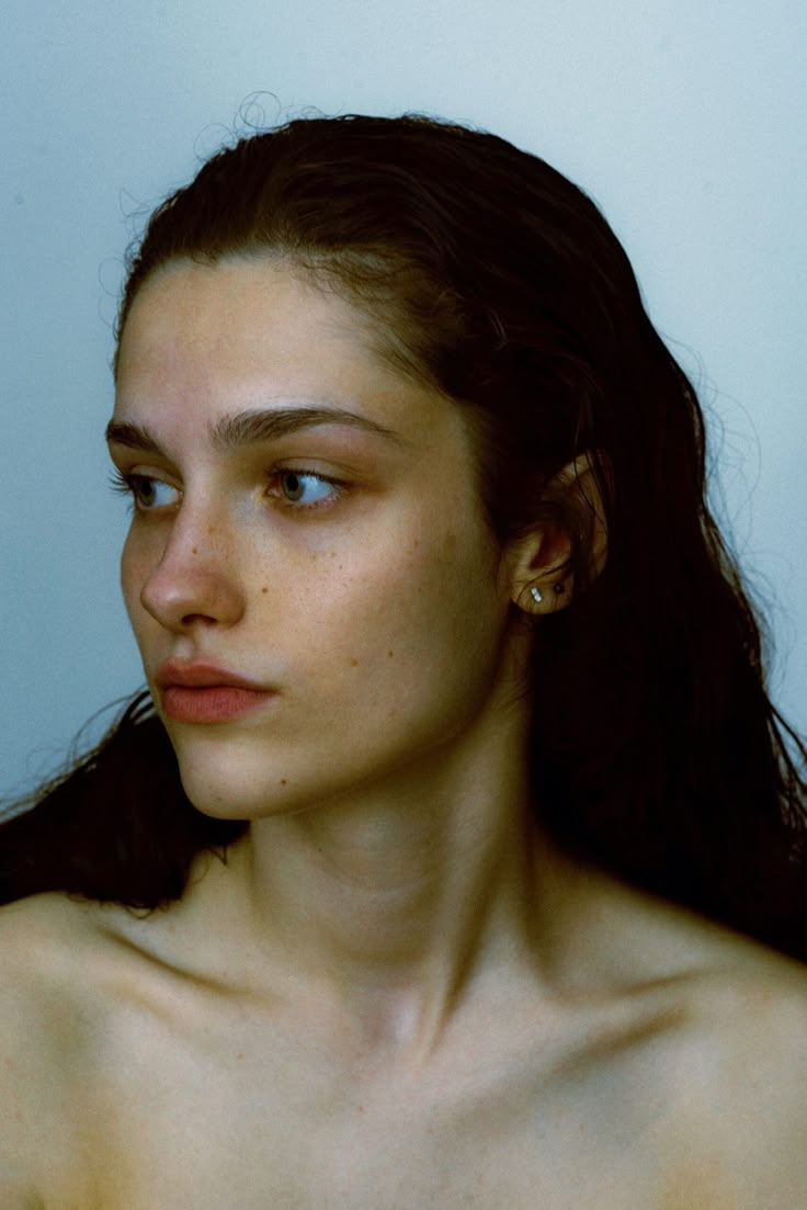 a woman with freckled hair and blue eyes looks off to the side in front of a white wall