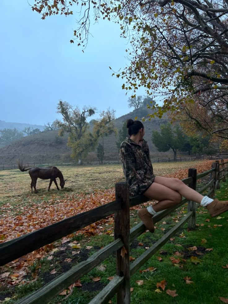 a woman sitting on a fence looking at an animal in the distance with leaves all around her