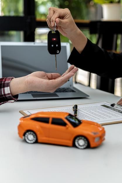 two people exchanging keys to an orange toy car on a table next to a laptop