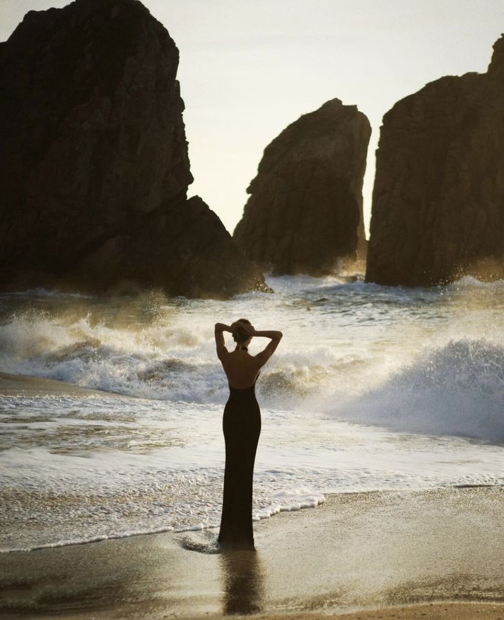 a woman standing on top of a sandy beach next to the ocean with rocks in the background