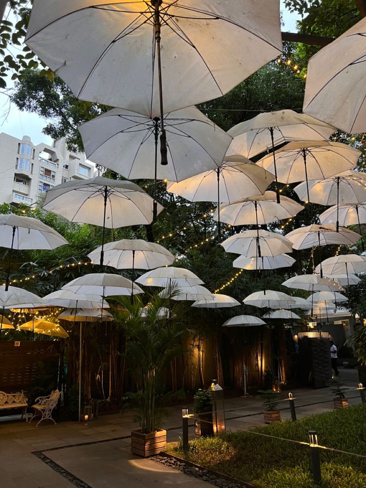 many white umbrellas are hanging from the ceiling in an outdoor area with lights and greenery