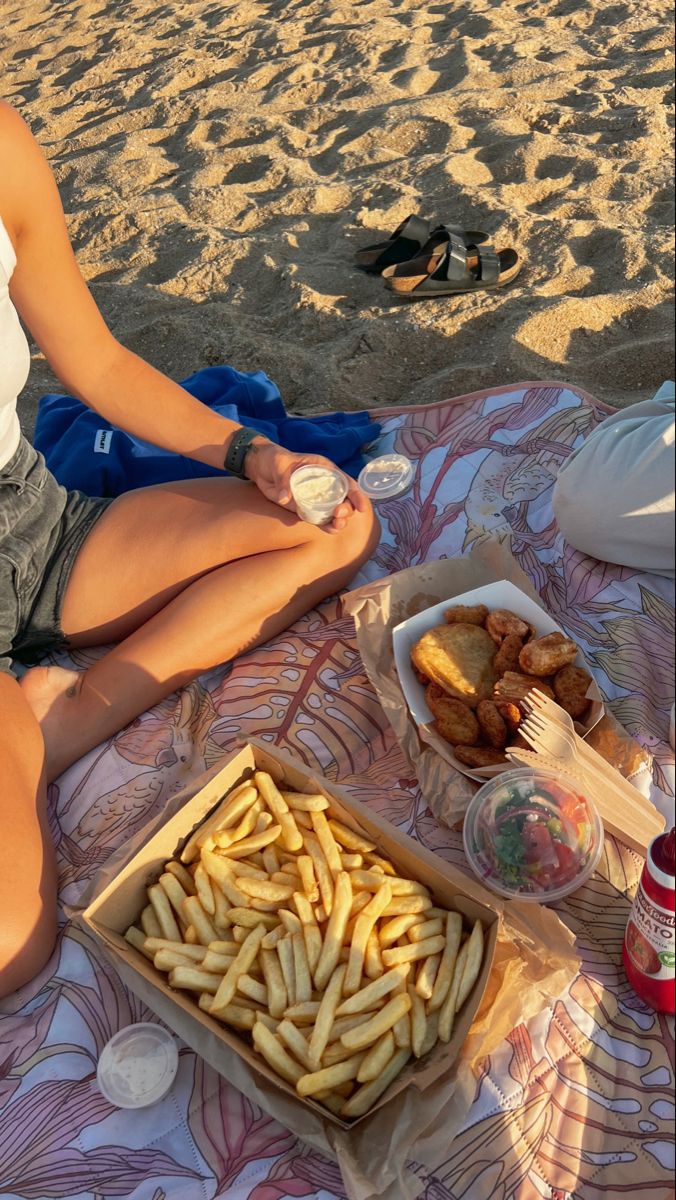 a woman is sitting on the beach with her feet crossed and food in front of her