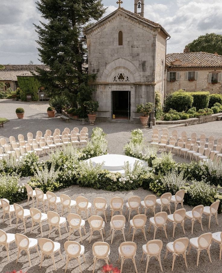 an outdoor ceremony setup with chairs and flowers in front of a church, surrounded by greenery