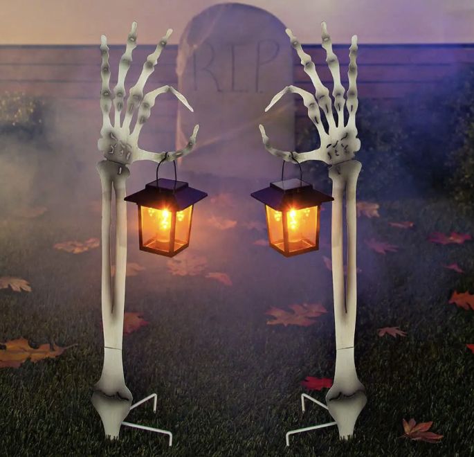 two halloween decorations made to look like skeleton hands holding a lantern in front of a tombstone