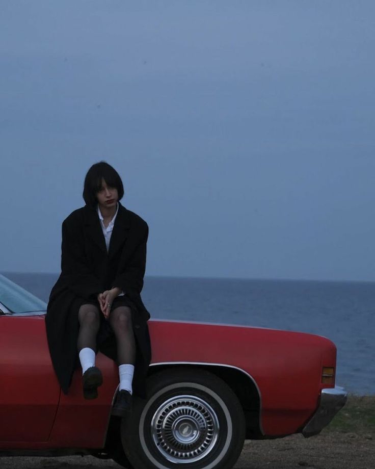 a woman sitting on top of a red car next to the ocean with her legs crossed