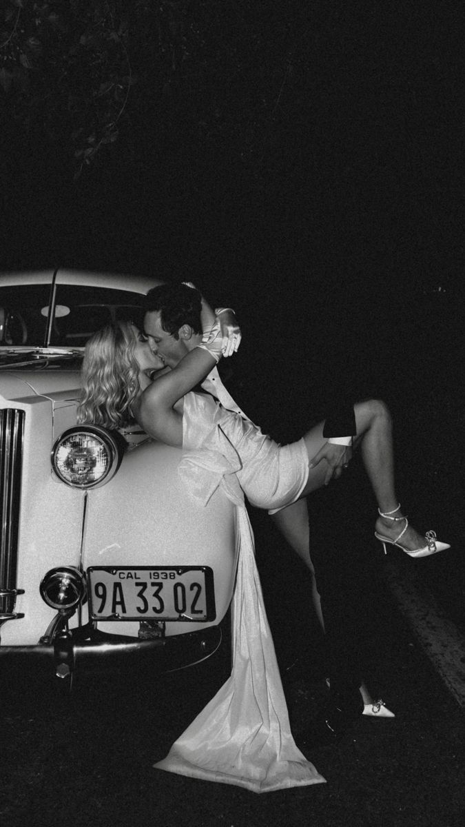 a man and woman kissing on the hood of a vintage car at night in black and white