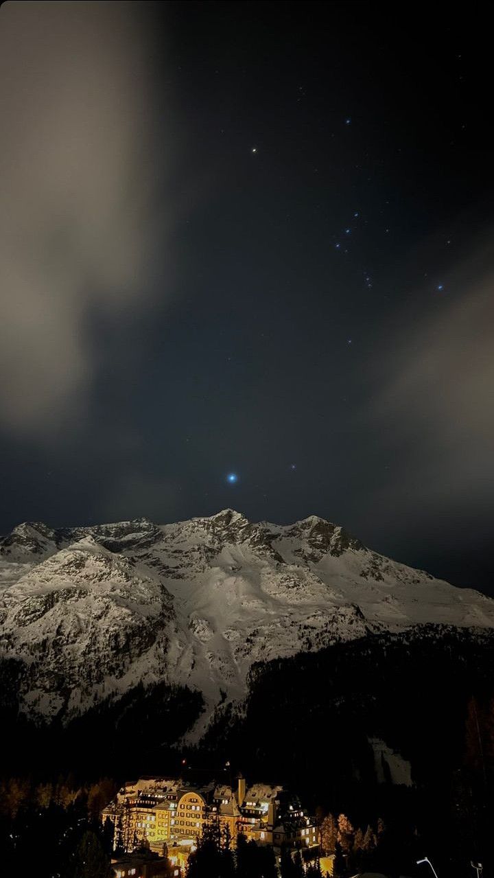 the night sky is lit up with stars and clouds above a snowy mountain range in the foreground