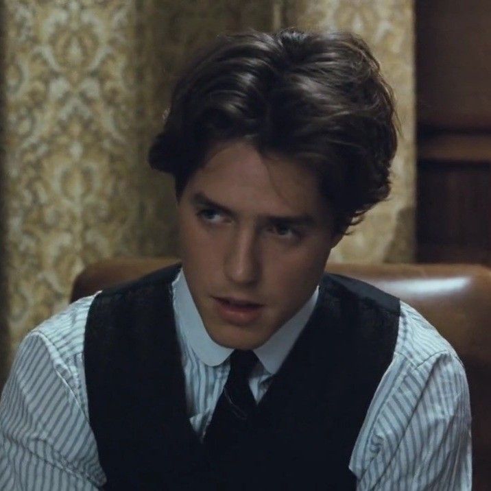 a young man wearing a vest and tie sitting in front of a brown leather chair