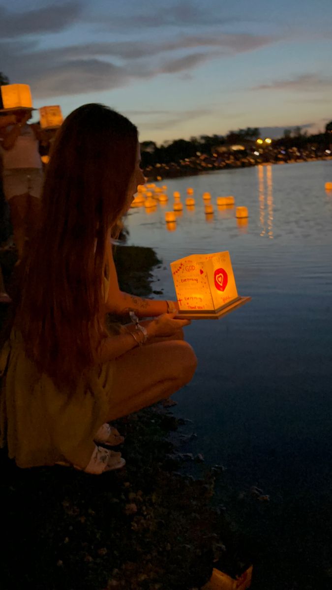 a woman sitting on the ground next to water holding a paper lantern in her hand