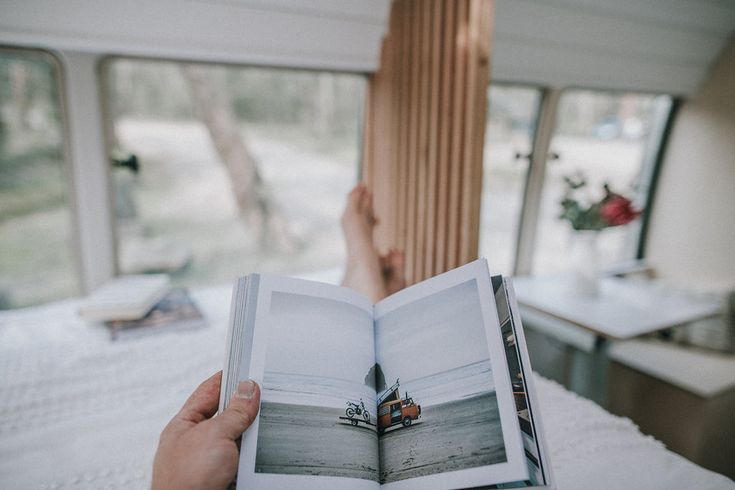 a person is holding an open book in their hand while sitting on a bed with windows