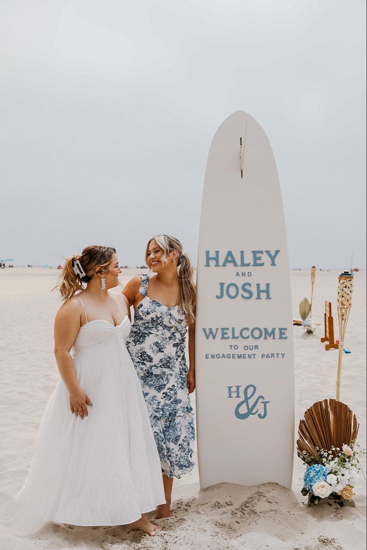 two women standing next to a welcome sign on the beach