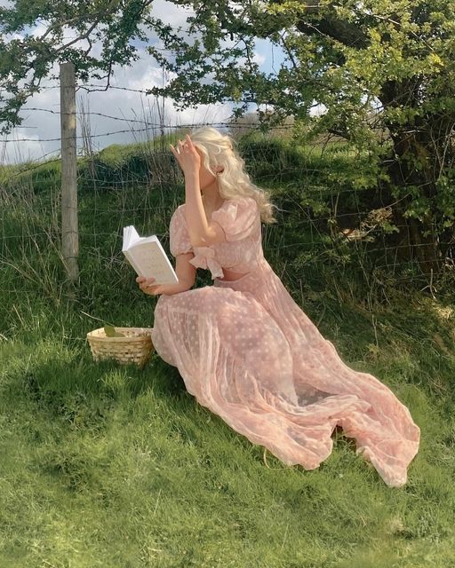 a woman in a pink dress sitting on the grass reading a book and leaning against a fence