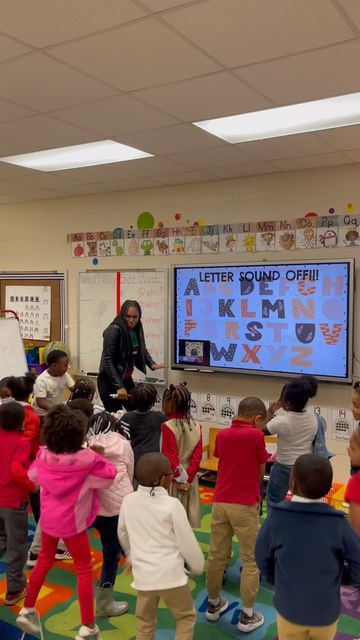 a teacher teaching children in a classroom with a large screen on the wall behind them