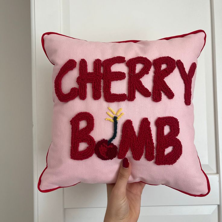 a hand holding up a pink pillow with the words cherry bomb written on it in red thread