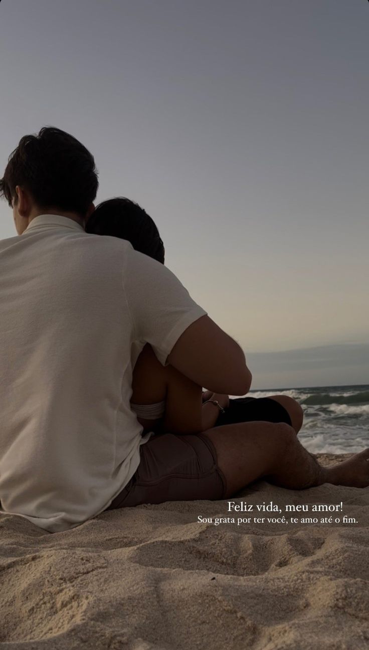 a man and woman sitting on the beach looking out at the ocean with their backs to each other