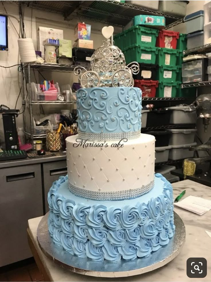 a three tiered cake with blue frosting and a tiara on top, sitting on a counter in a kitchen