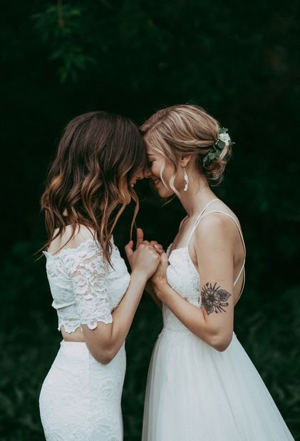 two beautiful women standing next to each other in front of green trees and bushes, one wearing a white dress