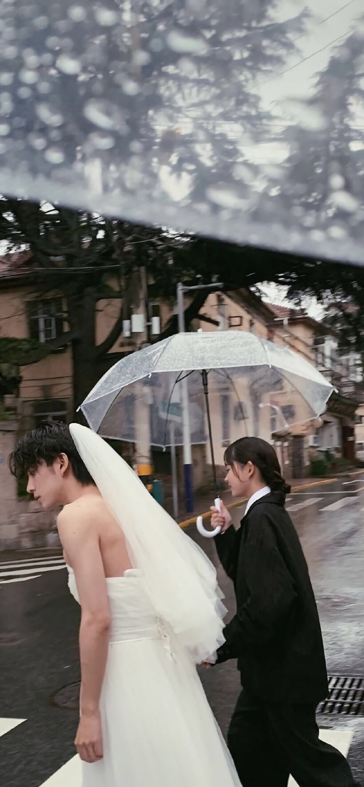 a bride and groom crossing the street in the rain with an umbrella over their heads