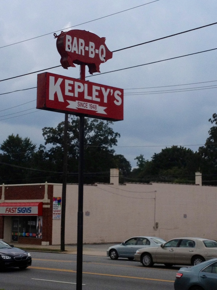 a bar - b - q sign on the corner of a street with cars parked in front of it