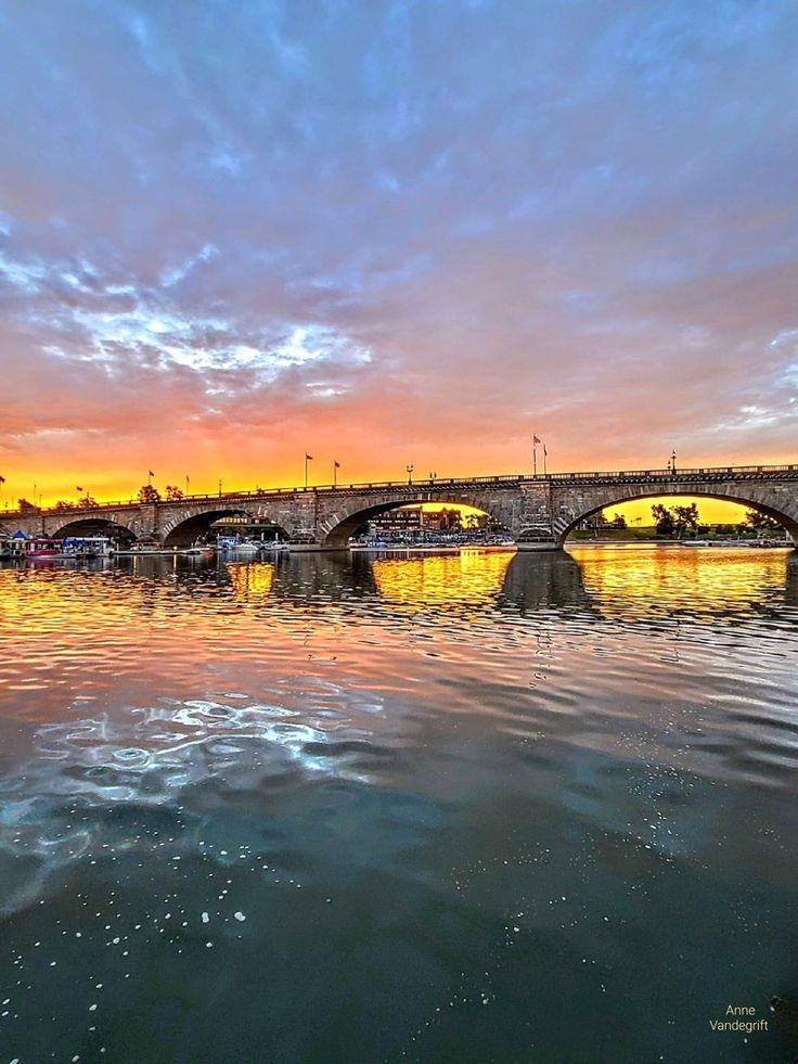 a bridge over water with boats on it and the sun setting in the sky above