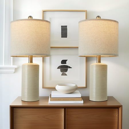 two lamps sitting on top of a wooden dresser next to a framed print and lamp shade