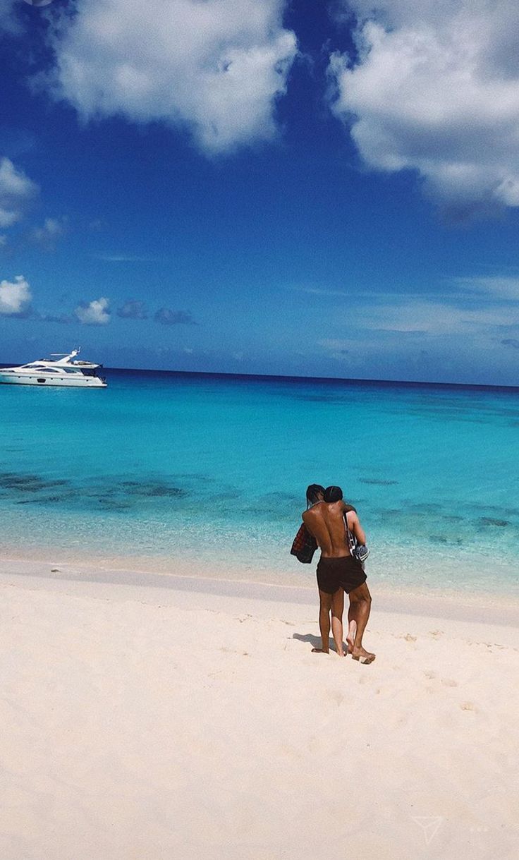 two people are standing on the beach with a boat in the background