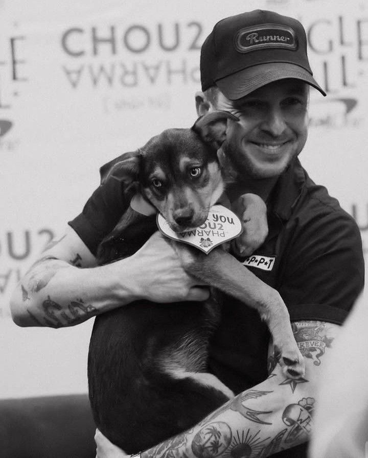 a man holding a dog in his arms while wearing a hat and tattoos on his arm