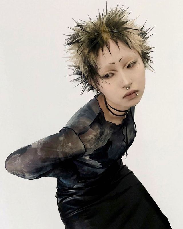 a woman with spiked hair and black dress posing in front of a white wall, her arms behind her head