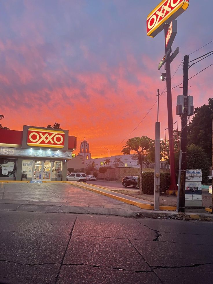 the sun is setting at an oxo store in front of some cars parked on the street