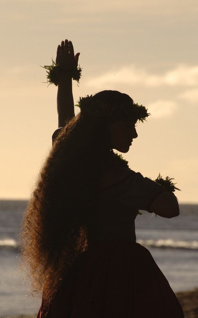 a woman with long hair standing in front of the ocean and holding her hands up