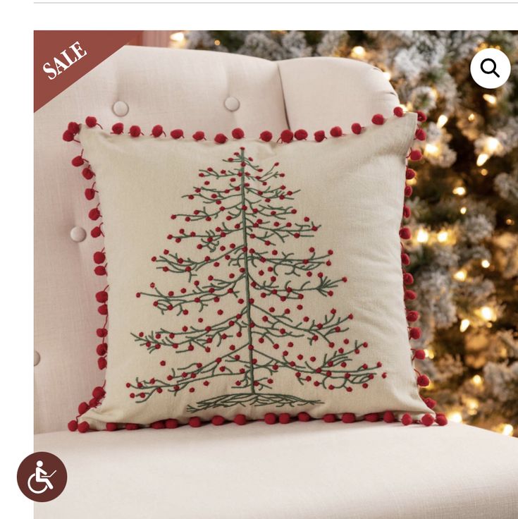 a christmas tree pillow on a white chair in front of a christmas tree with red pom - poms