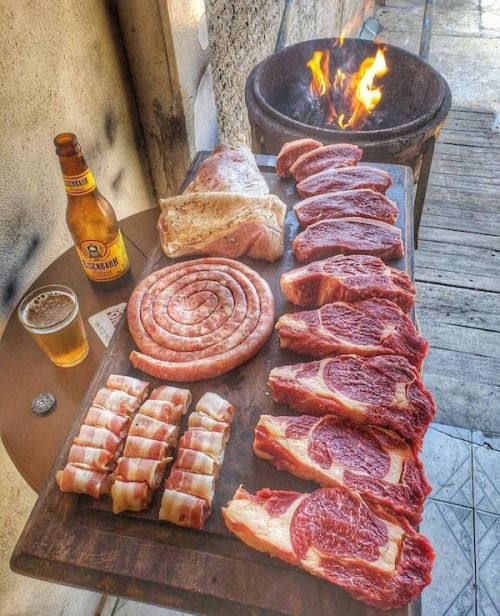 meats, sausages and beer sit on a table next to an open fire pit