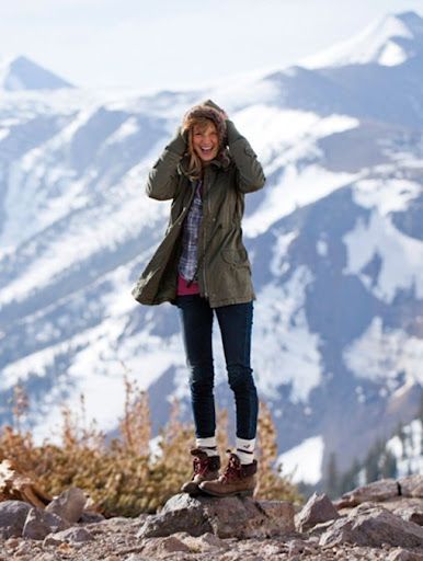 a woman standing on top of a mountain talking on a cell phone while wearing snow boots